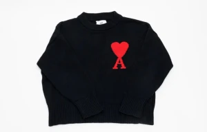 Svart stickad tröja från Ami - Svart stickad tröja från Ami med röd hjärtapplikation och bokstaven A på bröstet. Rund hals och ribbade muddar vid ärmslut och nederkant. Perfekt för dig som gillar stilrena detaljer och vill ha något som sticker ut lite extra.