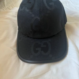 Svart keps från Gucci med GG-mönster - Snygg svart keps från Gucci med klassiskt GG-mönster ton-i-ton över hela kepsen. Justerbar rem baktill i skinn och böjd skärm. Tillverkad i slitstark bomull och polyester, perfekt för dig som vill ha en stilren och lyxig accessoar. Obs! Gucci lappen är inte på kepsen längre.