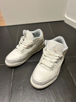 Nike Air Jordan 3 ivory craft  - Nike Air Jordan 3 Retro sneakers i vit och ljusgrå färg med klassisk Jumpman-logga på plösen och Nike Air på hälen. Skorna har snörning, rund tå och detaljer i skinn och mocka med mönstrad panel runt tån. Snygg siluett och platt sula, perfekt för dig som gillar streetwear.
