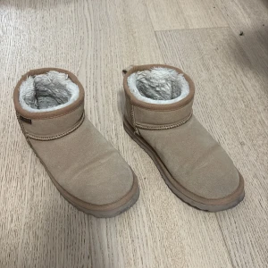 Beige boots med fluffigt foder - Mysiga beige boots med rund tå och platt sula, tillverkade i mocka med ett fluffigt vitt syntetfoder på insidan. Perfekta för kalla dagar och har en enkel, stilren design med dekorativ söm runt skaftet.