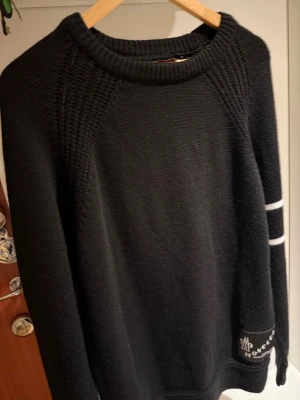 Svart stickad Moncler tröja med ränder - Svart stickad tröja från Moncler med två vita ränder på ena ärmen och klassiska Moncler-loggor. Rund hals och grovstickade detaljer vid axlarna. Perfekt för dig som gillar stilrena och exklusiva plagg med snygga detaljer.
