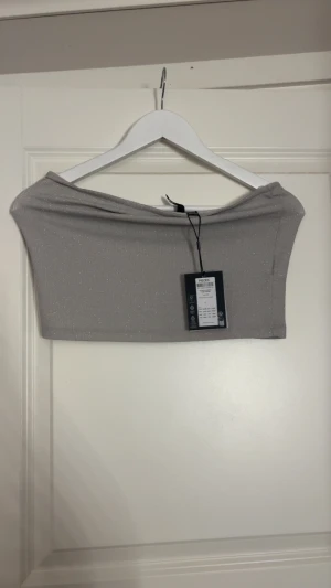 Glittrig silver tubtopp från Pieces - Säljer en snygg glittrig tubtopp i silver från Pieces. Toppen är axelbandslös och har en croppad passform som sitter tight. Perfekt för dig som vill sticka ut med lite extra glitter. Materialet är mjukt och stretchigt syntet.