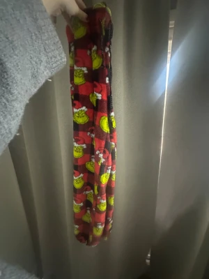 Grinch-mönstrade fleecebyxor - Mysiga fleecebyxor med rött och svart rutigt mönster och gröna Grinch-ansikten med tomteluva. Perfekta för chill hemma under vintern och julen. Byxorna har en loose passform och är supermjuka mot huden.