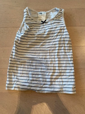 Randig linnetopp från H&M, 110/116 - Säljer en vit ärmlös linnetopp från H&M i storlek 110/116. Toppen har tunna svarta horisontella ränder och en liten mörkblå rosett vid halsringningen. Perfekt för varma dagar och enkel att matcha med jeans eller shorts.