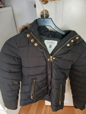 Svart pufferjacka med fuskpäls från Tindra - Svart pufferjacka från Tindra med guldiga dragkedjor och knappar. Jackan har huva med fuskpäls och två fickor framtill. Perfekt för kalla dagar och har en snygg, quiltad look. Passformen är normal och jackan är långärmad.