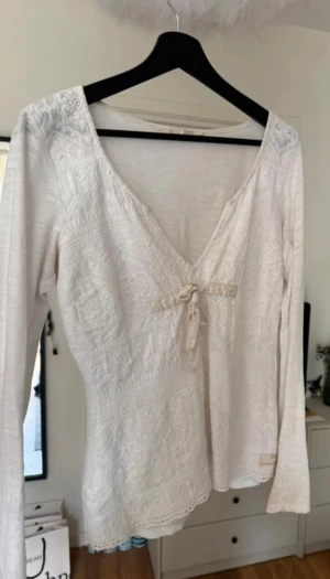 Vit Odd Molly blus med spets och knyt - Supersöt vit blus från Odd Molly med v-ringning, spetsdetaljer och ett litet knyt framtill. Långärmad och tillverkad i mjuk bomull med broderade mönster. Perfekt för dig som gillar romantisk och feminin stil.