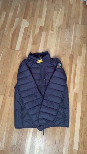 Mörkblå dunjacka från Parajumpers - Säljer en mörkblå dunjacka från Parajumpers med broderad patch på ärmen och gul detalj vid halsen. Jackan har quiltad design, dragkedja framtill och två sidofickor med dragkedja. Perfekt för kalla dagar och har en cool streetstil.