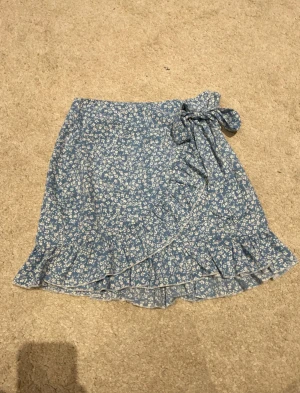 Blommig blå omlottkjol från SHEIN - Supersöt blå kortkjol från SHEIN med vitt blommönster. Kjolen har omlottmodell med knyt i sidan och volangkant längst ner, vilket ger en extra feminin touch. Perfekt för dig som gillar romantiska och trendiga plagg.