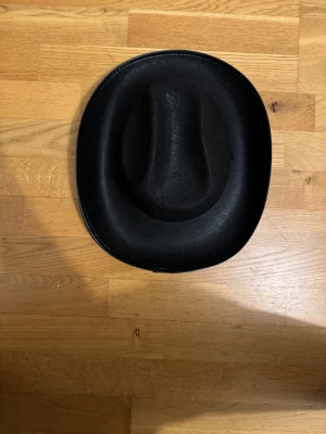 Svart cowboyhatt med snöre - Säljer en svart cowboyhatt med klassisk form och böjd brätte. Hatten har ett tunt snöre som kan knytas under hakan. Materialet är filtliknande och ger en cool westernkänsla. Perfekt för dig som vill sticka ut med en ikonisk accessoar.