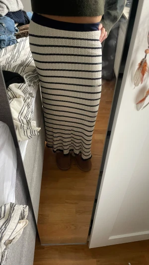 Randig långkjol från Atelier Rêve - Säljer en snygg långkjol från Atelier Rêve i storlek M. Kjolen är vit med mörkblå horisontella ränder och har en stickad struktur som ger en cool vibe. Midjan är elastisk och kjolen sitter tight över höfterna och faller rakt ner mot anklarna. Aldrig använd!!