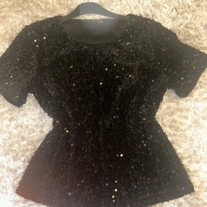 Svart paljettblus från Vero Moda - Säljer en svart blus från Vero Moda i storlek S, täckt med glittriga paljetter som ger en riktigt snygg och festlig vibe. Blusen har rund halsringning och korta ärmar, med en snygg passform. Perfekt för dig som vill sticka ut på nyår🎉