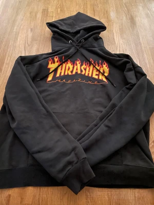 Svart Thrasher hoodie med flames - Svart hoodie från Thrasher med ikoniskt flames-tryck i gult och rött på bröstet. Tröjan har huva med snörning och en stor magficka. Mjuk och skön, perfekt för skate eller chill. Klassisk streetwear från San Francisco.