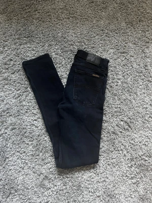Svarta skinny jeans från Nudie Jeans - Säljer ett par svarta skinny jeans från Nudie Jeans i storlek W28 L34. Jeansen har klassisk femficksdesign, svarta sömmar och Nudie Jeans logga på bakfickan. Materialet är stretchigt jeans och passformen är tight med smal siluett.