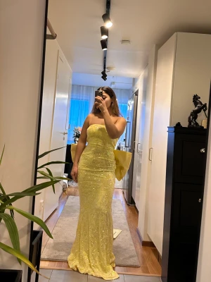 Gul glittrig balklänning med rosett - Säljer en lång, axelbandslös gul balklänning med glittrande paljetter och vackert mönster. Klänningen har en stor, dramatisk rosett och släp baktill som ger en riktigt wow-effekt. Perfekt för dig som vill sticka ut och glänsa på balen!