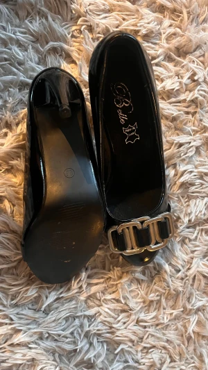 Svarta ballerinaskor med metallspänne - Snygga svarta ballerinaskor från Belle i lackat skinn med en chunky silverfärgad metallspänne framtill. Skorna har rund tå och klack, vilket gör dem både stilrena och trendiga. Perfekta för dig som gillar klassiska detaljer med en modern twist. Aldrig använda av mig 