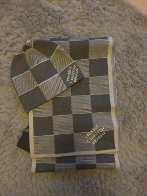 Louis vuttion halsduk och mössa  - Snygg mössa och halsduk i grått och vitt med klassiskt rutigt mönster. Stickad i mjukt material och har broderad text 'Marque L. Vuitton Deposee'. Perfekt för dig som vill ha en stilren accessoar med lyxig känsla.