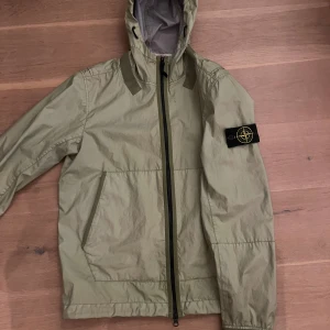 Stone Island jacka - Snygg grön vindjacka från Stone Island med huva och svart dragkedja. Jackan har klassisk patch på ärmen och är tillverkad i ett lätt och andningsbart material. Perfekt för dig som vill ha något stilrent och funktionellt.