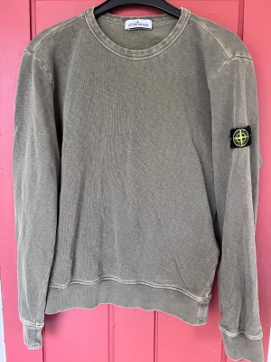 Grön sweatshirt från Stone Island Junior - Säljer en grön långärmad sweatshirt från Stone Island Juniori stl 156 med klassisk rund hals och ikonisk patch på vänster ärm. Tröjan är i mjuk bomull och har synliga sömmar för en avslappnad vibe. Perfekt för dig som gillar streetwear och snygga detaljer.