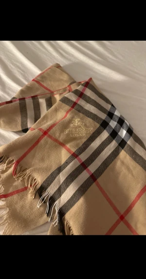 Beige rutig halsduk från Burberry - Klassisk beige halsduk från Burberry med ikoniskt rutigt mönster i svart, vitt och rött. Halsduken har fransar i kanterna och broderad Burberry-logga. Perfekt accessoar för att lyfta din outfit med en tidlös och lyxig känsla.