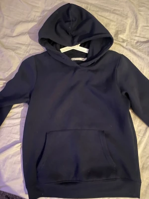 Mörkblå hoodie från 157, storlek 150 - Snygg mörkblå hoodie från 157 med klassisk huva och stor magficka framtill. Tröjan har långa ärmar och är gjord i mjuk bomullsmix som är skön att ha på sig. Perfekt för chill dagar och enkel att matcha med jeans eller mjukisbyxor.
