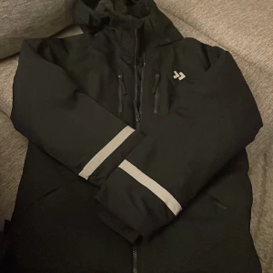 Svart vindjacka 157 Funk med reflex - Svart vindjacka från 157 Funk med huva, dragkedja och två bröstfickor med dragkedja. Jackan har reflexdetaljer på ärmar och rygg, samt logga på bröstet. Perfekt för blåsiga dagar och utomhusaktiviteter. 