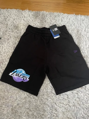 Svarta Los Angeles Lakers shorts XS - HELT NYA Svarta shorts från New Era med Los Angeles Lakers-logga i blått, vitt och lila på ena benet. Modellen har elastisk midja och är gjorda i mjuk bomull. Perfekta för dig som gillar basket och vill ha en chill streetwear-look. Pris kan Diskuteras!!