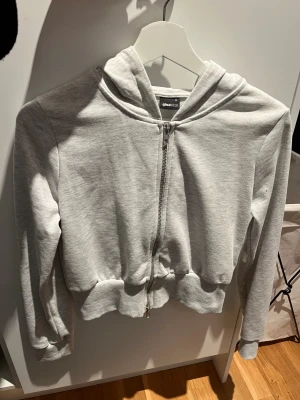 Ljusgrå hoodie från Gina Tricot - Säljer en ljusgrå croppad hoodie från Gina Tricot med dragkedja framtill och huva. Tröjan har långa ärmar, ribbade muddar och en croppad passform som sitter snyggt på kroppen. 