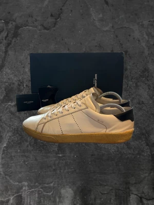 Saint Laurent SL/06 vita sneakers - Snygga Saint Laurent SL/06 sneakers i vitt skinn med beige gummisula och svart detalj på hälen. Klassisk rund tå, snörning och diskret perforering på sidan. Märkeslogga på plösen och yttersulan. Perfekt för dig som gillar stilrena och lyxiga sneakers.