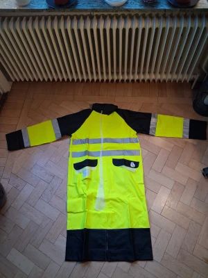Gul och svart regnjacka med reflex - Säljer en lång gul och svart regnjacka från LB med huva, stora fickor och breda reflexband över bröst, rygg och ärmar. Jackan har knappar framtill och detaljer i svart på fickorna samt logga. Perfekt när du vill synas och hålla dig torr.... ny pris ca 1500 kr