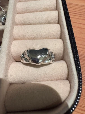 Silverring med hjärtformad detalj - Snygg silverfärgad ring med en stor hjärtformad detalj i mitten. Ringen har en blank yta och är lite chunky i stilen, perfekt för dig som gillar statement-smycken. Passar både som accessoar till din vardagslook eller när du vill sticka ut lite extra.