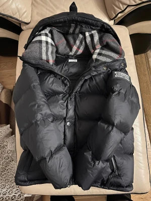 Svart jacka från Burberry - Svart jacka från Burberry med klassisk rutig huva i grått, vitt och rött. Perfekt för vintern. Pris går att diskutera! Etiketten är fortfarande på. 💯