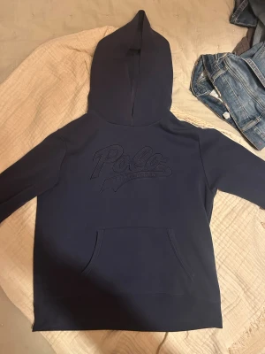 Mörkblå hoodie från Polo Ralph Lauren - Snygg mörkblå hoodie från Polo Ralph Lauren med stor broderad logga på bröstet. Tröjan har huva, känguruficka och långa ärmar. Tillverkad i mjuk bomull som är skön att ha på sig. Perfekt för dig som gillar klassisk streetstyle.