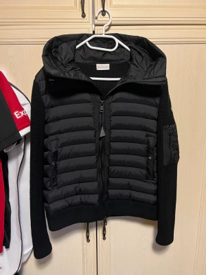 Svart Moncler Cardigan - Svart cardigan från Moncler med stickade ärmar och rygg, quiltad framsida och huva. Jackan har dragkedja framtill, fickor med dragkedja och Moncler-logga på ärmen. Snygg och sportig look med ribbade muddar och justerbar nederkant. Använd 4-5 gånger, mycket bra skick. Inga fel eller fläckar.