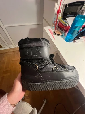 Svarta moon boots från Ella of Sweden - Säljer ett par svarta moon boots från Ella of Sweden. Skorna har en chunky platt sula, snörning med metallöglor och är tillverkade i syntetmaterial. Perfekta för vintern med sin vadderade och höga design som håller fötterna varma.