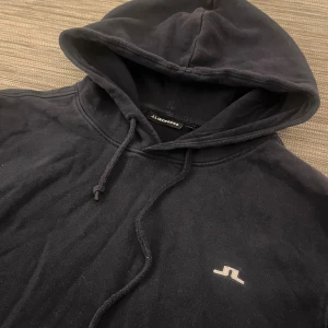 Marinblå hoodie från J.Lindeberg - Blå hoodie från J.Lindeberg med klassisk logga på bröstet. Tröjan har huva med snörning, magficka och ribbade muddar. Tillverkad i 100% ekologisk bomull, perfekt för chill dagar. Snygg och enkel design som funkar till det mesta.