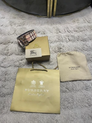 Burberry rutigt bälte med guldspänne - Snyggt bälte från Burberry med klassiskt rutigt mönster i beige, brunt och rött. Bältet har ett stort guldigt 'TB'-spänne och är gjort i skinn. Perfekt accessoar för att lyfta din outfit med en lyxig touch.
