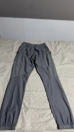 Grå träningsbyxor från Under Armour - Snygga grå träningsbyxor från Under Armour med elastisk midja och muddar vid bensluten. Byxorna har en tapered passform och är tillverkade i ett lätt syntetmaterial som andas. Perfekta för gymmet eller löprundan.