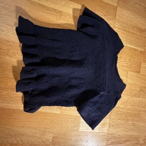 Svart blus med volangkant - Säljer en svart blus med korta ärmar och volangkant nertill. Toppen har en smockad struktur över hela kroppen och är tillverkad i ett mjukt tyg. Perfekt för dig som gillar en söt och trendig look.