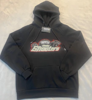 Svart Trapstar hoodie med broderi - Svart hoodie från Trapstar med fet broderad logga och texten 'Trapstar Shooters' på bröstet. Hoodien har snörning i huvan och en stor magficka. Materialet är mjukt och skönt, perfekt för chill dagar. Snygga detaljer med grått och rött broderi som sticker ut.