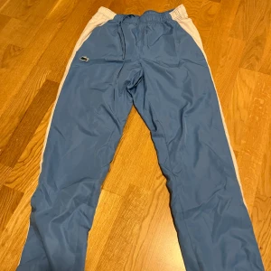 Blå träningsbyxor från Lacoste Sport - Snygga blå träningsbyxor från Lacoste Sport med vita sidopaneler och klassisk krokodillogga på benet. Byxorna har elastisk midja med snörning och är tillverkade i lätt syntetmaterial som andas. Regular fit för skön passform. Ny pris 1599kr