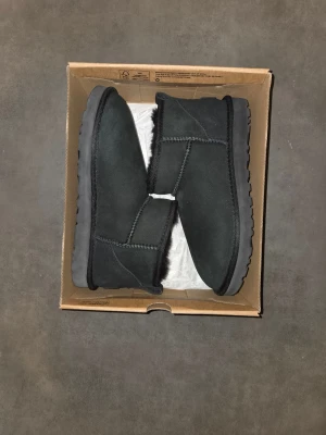Svarta boots från UGG - Säljer ett par klassiska svarta boots från UGG med låg skaft och rund tå. Skorna är i mocka med mjukt foder och har en platt sula. Perfekta för kyliga dagar och enkel att dra på tack vare hällan bak.