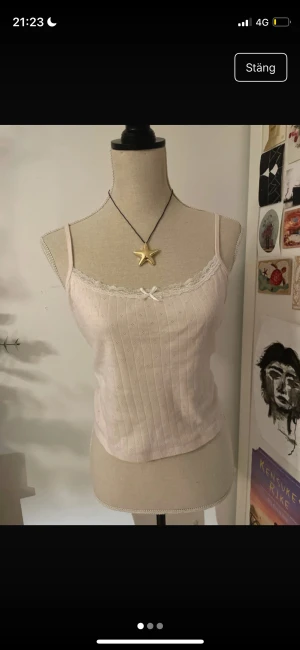 Ljusrosa ribbad linnetopp med spets - Supersöt ljusrosa linnetopp med smala axelband, ribbad struktur och små hålmönster. Toppen har spetskant och en liten rosett framtill för extra gullig vibe. Perfekt att styla med jeans eller kjol för en söt look.