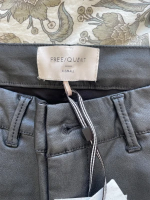 Svarta bootcut-byxor från Freequent - Snygga svarta byxor från Freequent i storlek XS. De har bootcut-ben, bälteshällor och klassisk midja. Materialet är slätt och har en lätt glansig finish som ger en modern vibe. Perfekta för dig som gillar stilrena och trendiga byxor.
