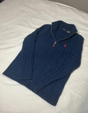 Marinblå stickad half zip Polo Ralph Lauren - Snygg marinblå stickad tröja från Polo Ralph Lauren med half zip och kabelstickat mönster. Tröjan har lång ärm, ribbade muddar och klassisk röd Polo-logga på bröstet. Perfekt för dig som gillar stilren och tidlös design.