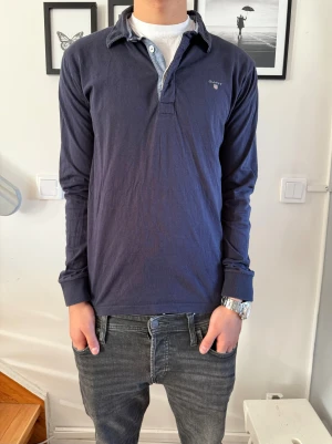 Marinblå pikétröja från GANT - Snygg marinblå långärmad pikétröja från GANT med klassisk krage och knappslå i ljusblått. Tröjan har diskret logga på bröstet och är gjord i skjort material. Perfekt för dig som gillar stilrena och tidlösa plagg. 