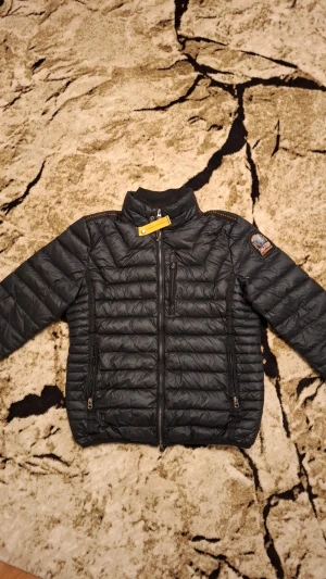 Svart dunjacka från Parajumpers - Snygg svart dunjacka från Parajumpers med broderad patch på ärmen och gul detalj vid halsen. Jackan har quiltad design, dragkedjor på fickorna och lång ärm. Insidan är grå och har praktiska fickor. Perfekt för dig som vill ha en lätt och stilren jacka.