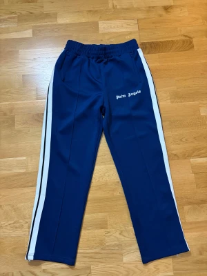 Blå Palm Angels trackpants med vita ränder - Snygga blå trackpants från Palm Angels med vita sidoremsor och logga broderad på framsidan. Byxorna har elastisk midja, raka ben och dragkedjor vid bensluten. Perfekt för en streetwear-look och riktigt sköna att bära.
