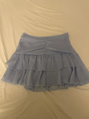 Ljusblå volangkjol  - Supersöt ljusblå kjol i storlek 34. Kjolen har flera lager med volanger och en snygg rynkning framtill. Tillverkad i ett lätt och luftigt syntetmaterial, med dragkedja i sidan för enkel påklädning.