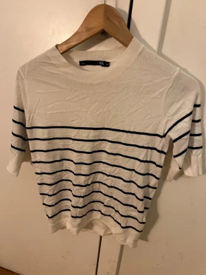 Vit randig stickad tröja 157 XS - Snygg vit stickad tröja från 157 i storlek XS med svarta horisontella ränder. Tröjan har rund hals och korta ärmar, perfekt för dig som gillar enkel och stilren design. Materialet är mjukt och lätt, vilket gör den skön att bära under vår och sommar.