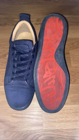 Mörkblå Louboutin sneakers mocka - Säljer ett par mörkblå sneakers från Louboutin i mjuk mocka med rund tå och platt sula. Skorna har snörning och signatur Louboutin-logga broderad på hälen. Yttersulan är röd med grafiskt mönster, och innersulan är beige. Perfekt för dig som gillar exklusiv streetstyle.